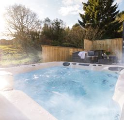 An outdoor hot tub with bubbling water next to a patio with wicker seating and a table with flowers at The Linhay Cadeleigh near Tiverton