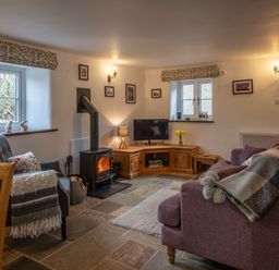 A living room with a wood-burning stove two windows a gray armchair and a purple sofa at Wavering in Compton Bishop near Axbridge