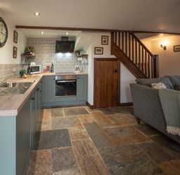 A kitchen with blue cabinets and a stone floor next to a gray sofa and wooden stairs at Crooks in Axbridge