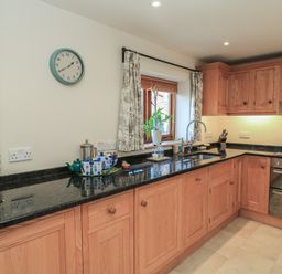 A kitchen with wooden cabinets black countertops a sink stove oven a window with curtains and a clock on the wall at Granary Cottage in Blackwell near Ilmington