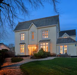 A large two-story house with lit windows and a gravel driveway surrounded by trees and lawn at Victoria Lodge in Kingsbridge