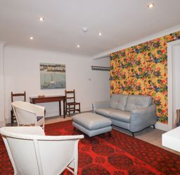 A living room with a gray sofa and ottoman two wicker chairs a red patterned rug a wooden table with two wooden chairs and a wall painting at Melvill Lower Deck in Falmouth