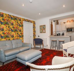 A living room with a grey sofa and ottoman two white wicker chairs and a blue armchair next to a kitchen with two stools with a floral wallpaper on one wall at Melvill Lower Deck in Falmouth