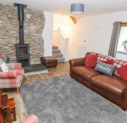 A living room with a stone fireplace wood stove a leather sofa with red cushions a plaid armchair a grey rug and stairs leading up at Y Beudy Crosswell Eglwyswrw near Newport Pembrokeshire