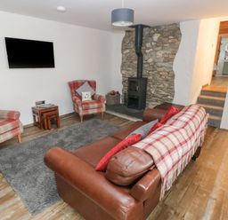 A living room with leather sofa, two plaid armchairs, a wall-mounted TV, a wood stove on a stone wall and a gray rug at Y Beudy in Crosswell, Eglwyswrw near Newport, Pembrokeshire