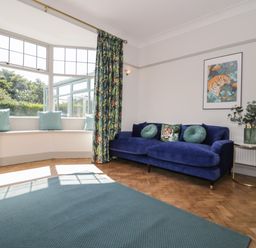 A living room with a blue sofa and round cushions a bay window with cushions floral curtains and a side table with plant at Monarfon in Amlwch