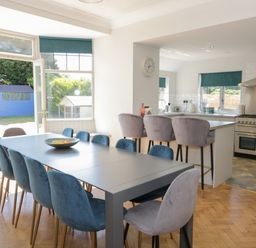 A dining area with a long table and blue chairs next to an open door and a kitchen with bar stools and a stove at Monarfon in Amlwch