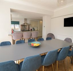 A dining area with a large table surrounded by blue chairs and three bar stools at a kitchen counter with beige cabinets at Monarfon in Amlwch