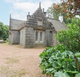A stone building with pointed roof and lattice windows surrounded by trees and plants at East Lodge Crathes Castle near Banchory
