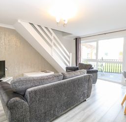 A living room with grey sofas a TV white tables a dining table and chairs and a staircase at 91 Waterside Park in Lowestoft