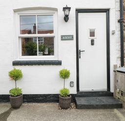 A white front door with a black frame next to a window with two potted plants in front at Ascot Cottage in Sheriff Hutton near Strensall