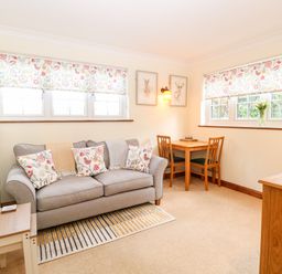 A sitting room with a gray sofa decorative cushions wooden table and chairs under windows at Hunters Lodge in Great Moulton near Long Stratton