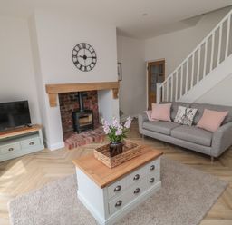 A living room with a gray sofa and pink cushions a TV on a stand a wood-burning stove and a coffee table with flowers at The Miners Cottage in Scremerston near Berwick-Upon-Tweed