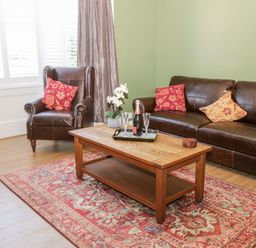 A sitting room with a brown leather sofa and armchair with red and beige cushions a wooden coffee table with a bottle and two glasses on a red patterned rug at Hazeldene in Sleights