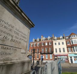 A street view with a stone monument engraved with text and row of mixed brick and white buildings with shops below a clear blue sky at The Kings Statue in Weymouth