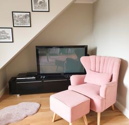 A pink armchair with matching footstool near a black TV stand with a television and framed pictures on the wall at The Seventeen in Bourton-On-The-Water