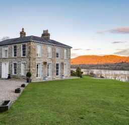 A stone house with multiple chimneys and white framed windows on a grassy lawn near a river with hills in the background at Silverholme Graythwaite near Hawkshead