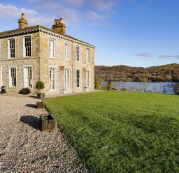 A stone house with large windows and chimneys next to a gravel path and a grassy lawn overlooking a lake and wooded hills at Silverholme in Graythwaite near Hawkshead