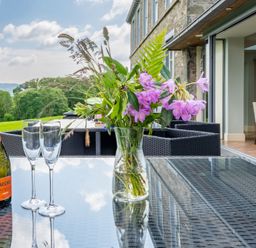 A glass table with a flower vase a bottle of prosecco and two glasses on a patio with outdoor chairs and a view of greenery at Silverholme Graythwaite near Hawkshead