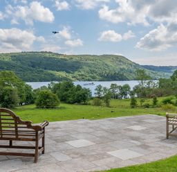 A patio with two wooden benches overlooking a lake and green hills with a plane in the sky at Silverholme in Graythwaite near Hawkshead