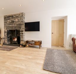 A living room with a stone fireplace a wall-mounted TV a wooden storage unit a brown leather sofa and wood stacked on shelves at Cooper's Rest in Dolwyddelan