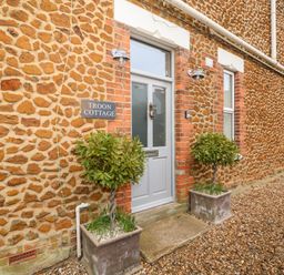Stone exterior wall with a grey door named Troon Cottage and two potted plants on a gravel path at Troon Cottage Hunstanton