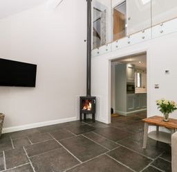 A living room with a wall mounted tv a small wood burning stove a wicker basket and a side table with flowers at Willow Barn in Shapwick