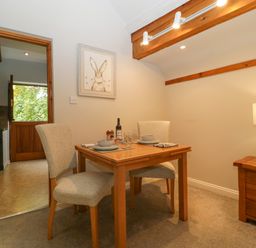 A dining area with a wooden table set for two and beige chairs next to a wooden nightstand with a lamp at The Coach House Apartment in Bredon