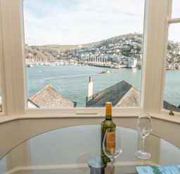 A glass table with a wine bottle and two wine glasses near windows overlooking a harbor with boats and houses at 4 Dartview in Dartmouth