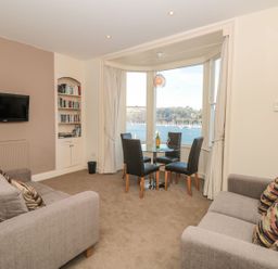 A living room with two grey sofas a wall-mounted television a built-in bookshelf and a dining table with four chairs by large windows overlooking a body of water at 4 Dartview in Dartmouth