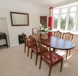 A dining room with a wooden table and six chairs near bay windows with red curtains at The Nook in Benllech