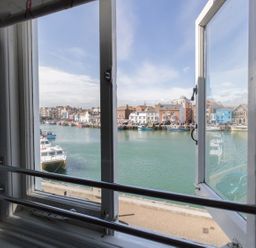 A view of boats docked in a harbor through an open window with buildings along the waterfront at Harbourside Cottage in Brewers Quay Harbour