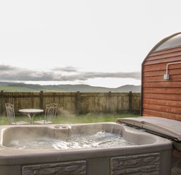 An outdoor hot tub next to a wooden cabin with a metal table and two chairs set on grass at Stapeley Pod in White Grit near Chirbury