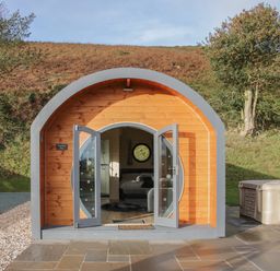 A curved wooden pod with open double doors showing a bed inside and a hot tub outside at Stapeley Pod in White Grit near Chirbury