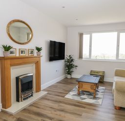 A living room with a wooden fireplace mantel, wall-mounted TV, beige sofa, wooden coffee table, indoor plants, and large window at The Croft Bungalow in Plumbland near Aspatria