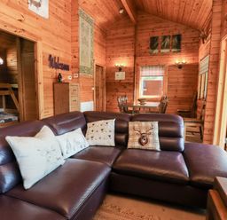 A wooden cabin interior with a brown leather sofa with cushions a dining table and chairs and an open door leading outside at Gisburn Forest Lodge Tosside near Wigglesworth