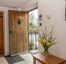 An entrance area with an open wooden door labeled Bluebell Cottage a wooden table with a glass vase of yellow flowers and a wall with a thermostat and a sign at Bluebell Cottage in Pembridge