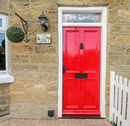 A red door with a black handle and mail slot on a stone wall with a hanging plant a lantern and a white picket fence at The Lodge in Stainsacre near Whitby