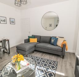 A living room with a gray sofa and colorful cushions a round mirror a glass coffee table with flowers and a dining table with metal chairs at Pebble Cove in Amble