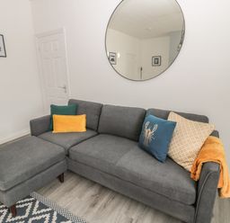 A grey sofa with colorful cushions and a blanket under a round mirror on the wall at Pebble Cove in Amble