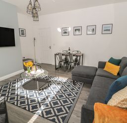 A living room with a gray sectional sofa with colored pillows a round glass coffee table on a patterned rug a dining table with four metal chairs and framed pictures on the wall at Pebble Cove in Amble