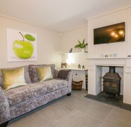 A living room with a gray sofa with green pillows a wood burning stove a wall mounted tv and a painting of a green apple at Apple Barn in Crantock