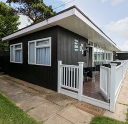 A black single-story house with white windows a white fenced porch with outdoor seating and a welcome mat at 98 Cherry Park in Chapel St Leonards