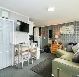 A small living area with a couch pillows a wooden chest of drawers a lamp a wall mirror a television two chairs and a kitchenette at 98 Cherry Park in Chapel St Leonards