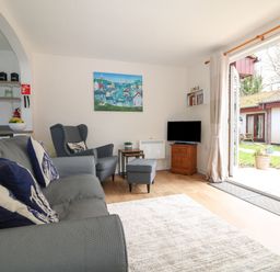 A living room with a grey sofa and armchair a small TV on a wooden stand and open doors to a garden at Miles Retreat in Hayle