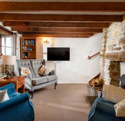 A living room with wooden beams a stone fireplace two blue armchairs a grey sofa a wooden side table a lamp bookshelves and a wall-mounted TV at Ash Cottage in Warbstow