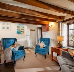 A sitting room with blue armchairs wooden beams stone wall and a window with flowers at Ash Cottage in Warbstow