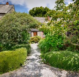 A garden path with neatly trimmed bushes and a large arching shrub leading to a stone house at Pond Meadow in Warbstow