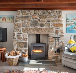 A living room with a stone fireplace and wood burner two paintings on the wall a television on wooden tables a basket of firewood a grey armchair with yellow floral pillows at Pond Meadow in Warbstow