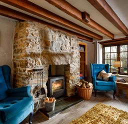 A living room with blue armchairs a stone fireplace a basket with firewood and a window at Little Leighs in Warbstow